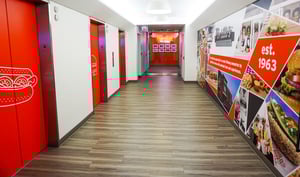 Portillo's office entrance with vinyl hot dog graphics on the elevators, a mural of their offerings on the right wall, and custom lit signage in the distance.
